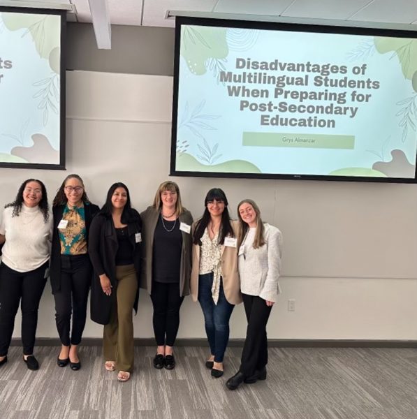 A group of six women pose in front of a slideshow presentation.